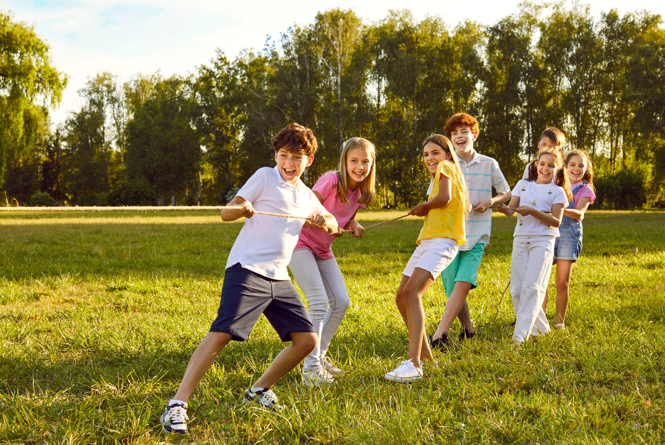 Happy kids playing games and having fun in summer camp on vacation. Team of children playing tug of war, standing on green field and pulling rope all together. Summer, fun, activity, teamwork concept